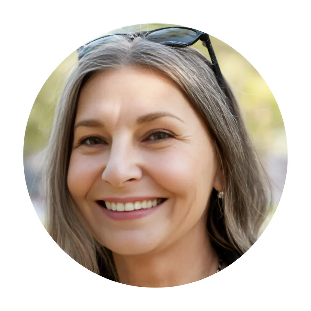 A close-up headshot of a smiling middle-aged woman with shoulder-length, straight hair that is graying. She has dark sunglasses resting on top of her head, is wearing small hoop earrings, a delicate silver and gold chain necklace, a white top, and a black leather jacket. She looks slightly to the side with a warm, cheerful expression against a softly blurred, natural outdoor background.