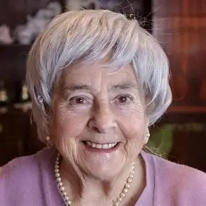 A close-up headshot of a smiling elderly woman with short, styled silver-gray hair. She is wearing small pearl earrings, a pearl necklace, and a lavender pink top. She looks directly at the camera with a warm expression, displaying visible smile lines and wrinkles on her face.
