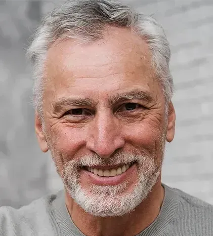 A close-up headshot of a smiling older man with short, graying hair and a neatly trimmed gray beard and mustache. He is looking directly at the camera with warm brown eyes, showing prominent smile lines and wrinkles around his eyes and forehead, and wearing a gray crew-neck shirt against a soft-focus gray background.
